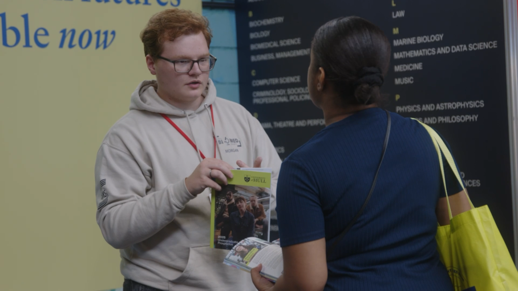 A young man wearing glasses and a beige hoodie with a name tag reading ‘Morgan’ engages with a woman at an event. He holds a prospectus from the University of Hull, while the woman, carrying a yellow tote bag, listens attentively. Educational posters are visible in the background. Taken from the Prism Report by Prospectus Plus.