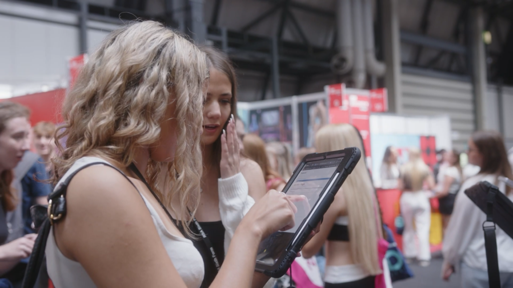 Two young women at an event, with one holding and interacting with a tablet while the other watches intently. The scene is busy with people and colourful booths in the background, indicating an exhibition or fair. Taken from the Prism Report by Prospectus Plus.