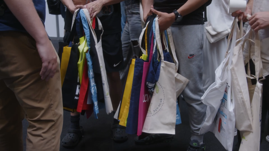 Close-up of individuals holding multiple tote bags of various colours and designs, suggesting they have collected them from an event or exhibition. The image focuses on their hands and the bags, with casual attire including trousers, shorts, and trainers visible. Taken from the Prism Report by Prospectus Plus.