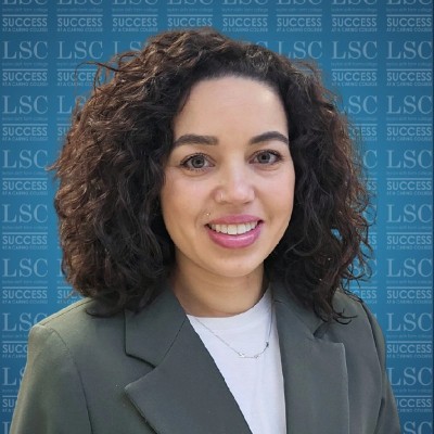 Woman with curly dark hair and a green blazer smiling in front of a branded Leyton Sixth Form College backdrop featuring the LSC logo and the slogan “Success at a Caring College.”