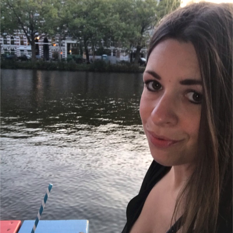 A woman with long brown hair and subtle makeup smiles slightly while sitting beside a calm river. Trees and buildings line the opposite bank in the background. A blue and white striped straw and colourful table are partially visible in the foreground.