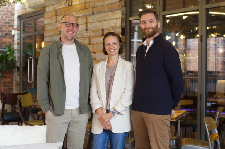 Three people standing together and smiling for a photo in a modern indoor venue with wood-panelled walls and glass doors.
