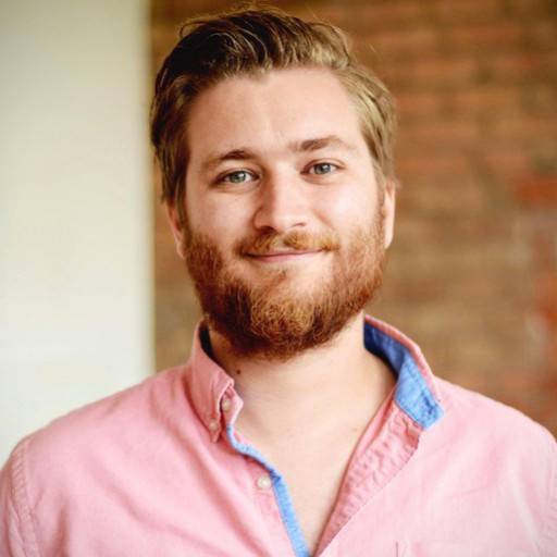 A photographic portrait of Nathan Monk, a white male, with medium length blonde hair, swept back, with a medium length beard. He is wearting a bink shirt, set against a brick wall backdrop.